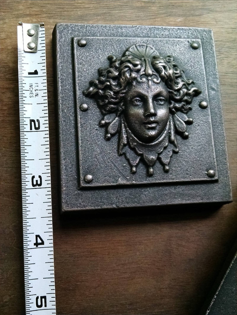 Pu&ograve; includere: Una placca da parete in metallo argentato con una scultura a rilievo di un viso di donna. La donna ha i capelli lunghi e ricci e indossa una corona. La placca &egrave; larga circa 10 cm e alta 12,5 cm.
