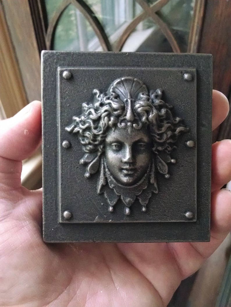 Pu&ograve; includere: Una placca di metallo grigio con una scultura a rilievo di un viso di donna. La donna ha i capelli lunghi e ricci e indossa una corona con una conchiglia in cima. La placca ha un bordo decorativo con piccoli punti.