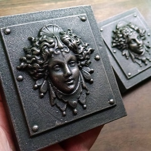 Pu&ograve; includere: Due placche quadrate in metallo con un designo in rilievo dettagliato di un viso di donna con capelli fluenti e gioielli decorativi. Le placche hanno una finitura scura invecchiata e sono accentate da borchie argentate.
