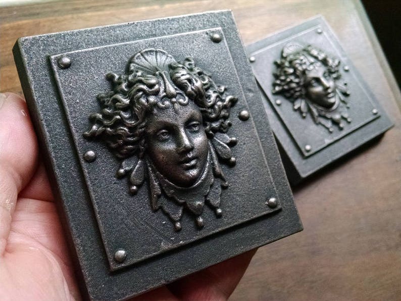 Pu&ograve; includere: Due placche quadrate in metallo con un designo in rilievo dettagliato di un viso di donna con capelli fluenti e gioielli decorativi. Le placche hanno una finitura scura invecchiata e sono accentate da borchie argentate.