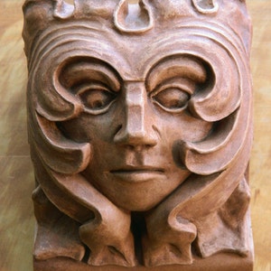 Könnte beinhalten: Eine braune Terrakotta-Skulptur eines Gesichts mit aufwendigen Details. Das Gesicht ist von einem dekorativen Rand aus Blättern und Spiralen umrahmt.