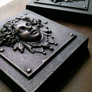 Pu&ograve; includere: Due placche quadrate in metallo per parete con sculture in rilievo dettagliate e tridimensionali di volti. I volti sono scolpiti in stile classico con capelli lunghi e fluenti e tratti delicati. Le placche hanno una finitura metallica scura e invecchiata.