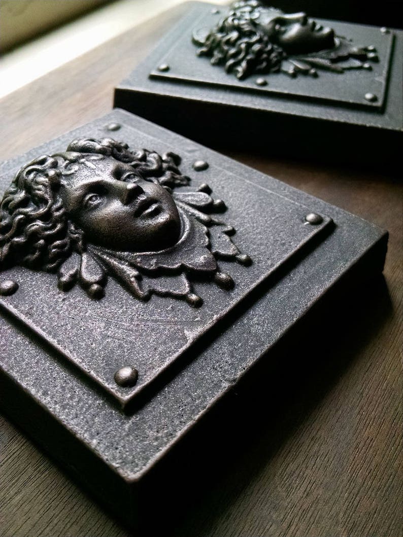 Pu&ograve; includere: Due placche quadrate in metallo per parete con sculture in rilievo dettagliate e tridimensionali di volti. I volti sono scolpiti in stile classico con capelli lunghi e fluenti e tratti delicati. Le placche hanno una finitura metallica scura e invecchiata.