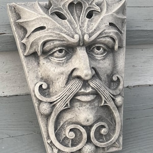 May include: A gray stone sculpture of a Green Man's face with a leafy crown and a detailed beard. The sculpture is mounted on a wall.