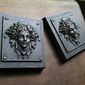 Pu&ograve; includere: Due placche quadrate in metallo per parete con un design in rilievo dettagliato di un viso di donna con capelli fluenti. Le placche sono di colore grigio scuro con un riflesso metallico.