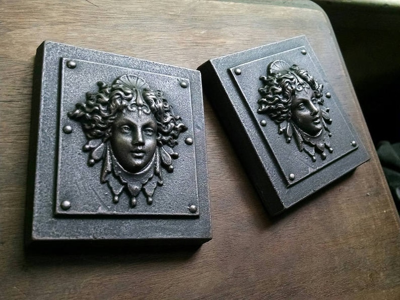 Pu&ograve; includere: Due placche quadrate in metallo per parete con un design in rilievo dettagliato di un viso di donna con capelli fluenti. Le placche sono di colore grigio scuro con un riflesso metallico.