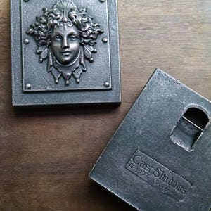 Pu&ograve; includere: Due placche da parete in metallo, una con un rilievo in rilievo di un viso femminile e l'altra con il testo "Cast Shadows 1995 & Co."