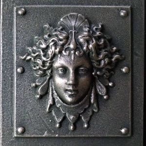 Pu&ograve; includere: Una placca in metallo color argento con una scultura a rilievo di un viso di donna. La donna ha capelli lunghi e fluenti e un copricapo delicato e ornato. La placca ha un aspetto testurizzato e invecchiato.