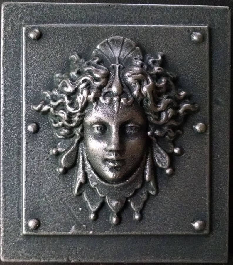 Pu&ograve; includere: Una placca in metallo color argento con una scultura a rilievo di un viso di donna. La donna ha capelli lunghi e fluenti e un copricapo delicato e ornato. La placca ha un aspetto testurizzato e invecchiato.