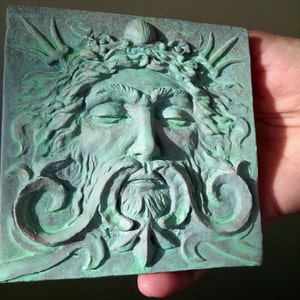 May include: A green ceramic wall plaque with a detailed relief sculpture of a man's face with closed eyes and a beard. The face is surrounded by stylized floral and leaf designs.