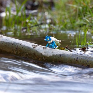 May include: A blue and white figurine of a creature with large eyes and a fluffy collar sits on a log in a stream. The water flows around the log, and green grass is visible in the background. The figurine has a small, white, pearl-like structure on its back.