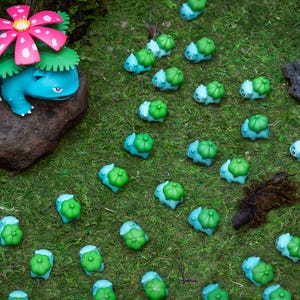 May include: A collection of blue and green figurines resembling a popular character, scattered on a bed of green moss. One larger figurine with a pink and white flower sits atop a rock. The scene evokes a whimsical, fantasy theme.