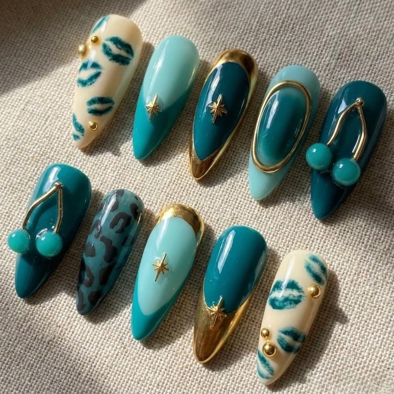 May include: A set of ten almond-shaped press-on nails featuring a variety of designs including cream nails with green lip prints, solid teal nails, teal nails with gold star charms, teal nails with gold cherry charms, and teal leopard print patterns.
