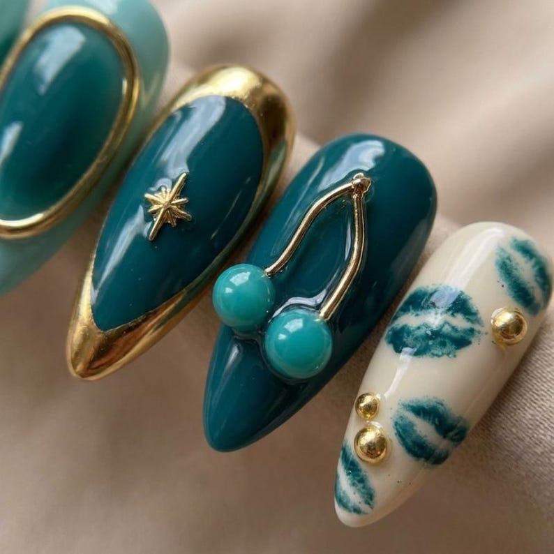 May include: A set of four almond-shaped press-on nails featuring a deep teal glossy finish. One nail has a gold star charm, another features a gold-toned cherry charm with teal beads, and the final nail is cream-colored with teal lip prints and gold studs.