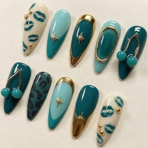 May include: A set of ten almond-shaped press-on nails featuring a variety of designs including cream nails with green lip prints and gold studs, teal nails with gold star charms, teal nails with gold frames, and teal nails with gold cherry charms.
