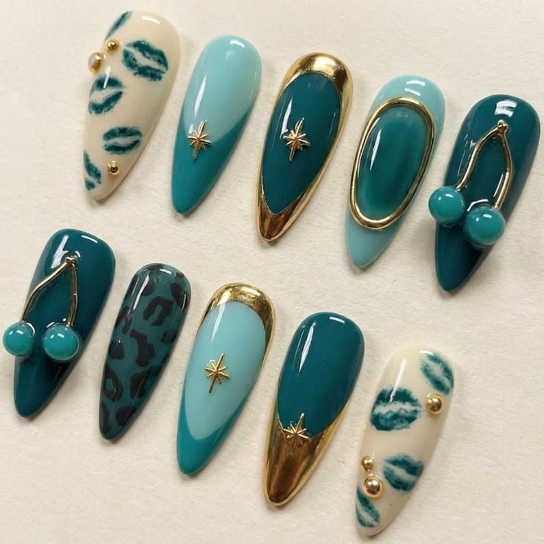 May include: A set of ten almond-shaped press-on nails featuring a variety of designs including cream nails with green lip prints and gold studs, teal nails with gold star charms, teal nails with gold frames, and teal nails with gold cherry charms.