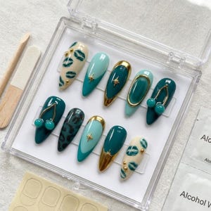 May include: A set of ten press-on nails in a clear plastic case, featuring almond-shaped tips in shades of teal, mint green, and cream. Designs include gold star charms, gold-rimmed accents, green leopard print, lip patterns, and dangling charms.