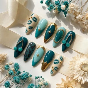 May include: A set of ten almond-shaped press-on nails featuring a color palette of cream, light teal, and dark teal. Designs include dark teal lip prints on cream, gold star charms, gold metallic tips, a gold oval frame, and 3D teal cherry charms.