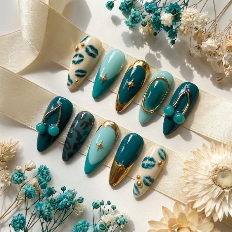 May include: A set of ten almond-shaped press-on nails featuring a color palette of cream, light teal, and dark teal. Designs include dark teal lip prints on cream, gold star charms, gold metallic tips, a gold oval frame, and 3D teal cherry charms.