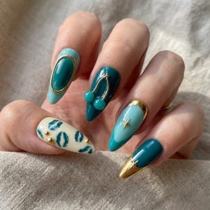 May include: Almond-shaped nails featuring a mix of teal and light blue polish. Designs include a gold oval frame, a 3D cherry charm, a starburst accent, gold foil tips, and a cream-colored nail with teal lip prints and small gold studs.
