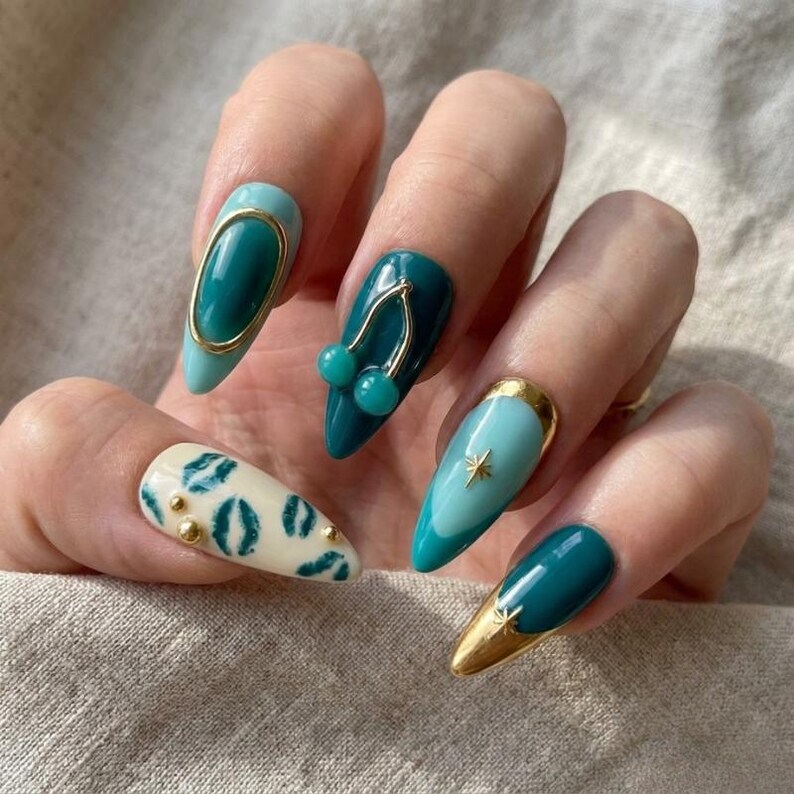 May include: Almond-shaped nails featuring a mix of teal and light blue polish. Designs include a gold oval frame, a 3D cherry charm, a starburst accent, gold foil tips, and a cream-colored nail with teal lip prints and small gold studs.