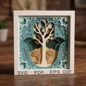 May include: A square white frame containing a multi-layered paper cut-out art piece featuring a central tree and two flowers within a hamsa shape. The design uses shades of light blue, dark green, and beige. The text SVG PDF EPS DXF is printed.