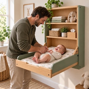 Può includere: Un fasciatoio a parete in legno con pannelli laterali color verde salvia e finitura in legno chiaro. L'unità è dotata di tre ripiani aperti contenenti biancheria piegata, un cesto intrecciato, una pianta in vaso, un portafazzoletti e un coniglio di peluche.