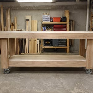 Könnte beinhalten: Eine rechteckige Werkbank aus Holz mit einer dicken, hellen Holzplatte und einer passenden unteren Ablagefläche. Die Konstruktion ruht auf vier stabilen Beinen, die jeweils mit einer schwenkbaren Metallrolle für Mobilität ausgestattet sind.