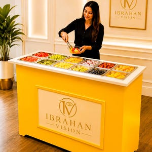 Può includere: Una postazione di servizio rettangolare giallo brillante con un piano di lavoro bianco e dodici vaschette in acciaio inossidabile riempite di frutta fresca assortita. Il pannello frontale mostra il logo e il testo dorati IBRAHAN VISION.