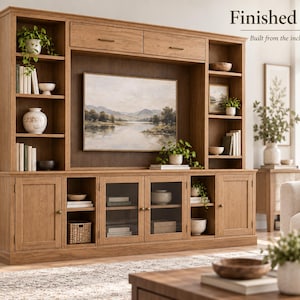 May include: A large light brown wooden entertainment centre featuring open shelving, two upper drawers, and lower cabinets with solid and glass-panelled doors. The unit displays books, potted plants, bowls, and a landscape painting in the centre.