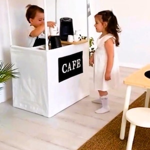May include: A white play cafe stand with a black and white striped awning. The stand has a black sign that says "CAFE" in white letters. A child is behind the counter, and another child is standing in front of the stand.