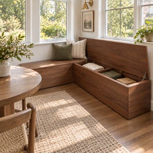 Può includere: Una panca contenitore in legno a forma di L con finitura in noce scuro, dotata di un coperchio incernierato che si apre per rivelare scomparti interni. La panca è posizionata in un angolo sotto le finestre e include un cuscino verde oliva.