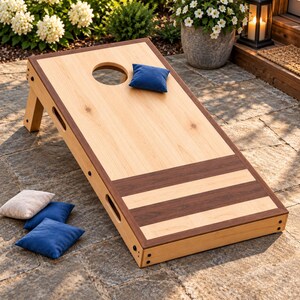 Könnte beinhalten: Ein Cornhole-Spielbrett aus Holz mit einer hellen Naturholzoberfläche, einem kreisförmigen Loch, dunklen Holzkanten und zwei horizontalen dunklen Holzstreifen an der Basis. Ein blauer Sitzsack liegt auf dem Brett, während zwei weitere blaue und ein beiger Sitzsack daneben liegen.