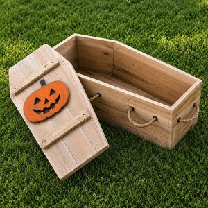 May include: A light brown wooden coffin-shaped box with a removable lid featuring a carved orange jack-o'-lantern pumpkin. The box includes thick rope handles attached to the sides with metal hardware, showcasing a natural wood grain finish.