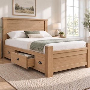 Op de afbeelding: Een bedframe van licht eikenhout met een hoog hoofdeinde en geïntegreerde opberglades aan de onderkant. Het bed is opgemaakt met wit beddengoed, een saliegroene sprei en twee kussens. De lades zijn voorzien van kleine, donkere metalen grepen.