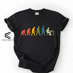Peut inclure: Un t-shirt noir &agrave; manches courtes arborant un graphique color&eacute; de l'&eacute;volution humaine, allant d'un primate &agrave; une personne assise &agrave; un bureau avec un ordinateur portable. Le motif inclut le texte CRAFTYNOVA sur le c&ocirc;t&eacute; gauche du tissu.
