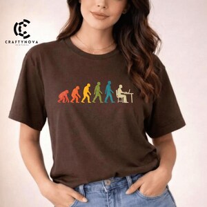 Peut inclure: Un t-shirt &agrave; manches courtes marron fonc&eacute; avec une impression graphique color&eacute;e repr&eacute;sentant la s&eacute;quence de l'&eacute;volution humaine. Le motif montre la progression d'un primate vers un humain assis &agrave; un bureau avec un ordinateur, en rouge, orange, jaune, vert et bleu.