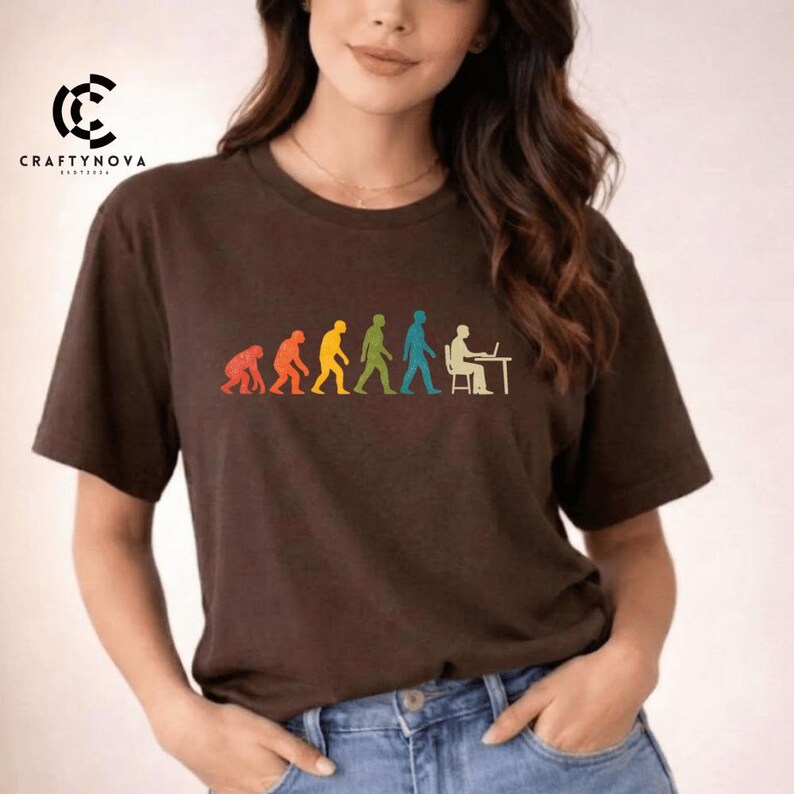 Peut inclure: Un t-shirt &agrave; manches courtes marron fonc&eacute; avec une impression graphique color&eacute;e repr&eacute;sentant la s&eacute;quence de l'&eacute;volution humaine. Le motif montre la progression d'un primate vers un humain assis &agrave; un bureau avec un ordinateur, en rouge, orange, jaune, vert et bleu.