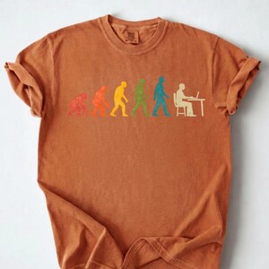 Peut inclure: Un t-shirt &agrave; manches courtes de couleur rouille avec une impression graphique de l'&eacute;volution humaine dans un style vieilli. Le motif montre une s&eacute;quence de silhouettes progressant d'un primate &agrave; une personne assise &agrave; un bureau avec un ordinateur.