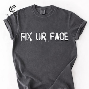 May include: Dark gray t-shirt with the text "FIX UR FACE" in white, with a dripping effect. The shirt has rolled-up sleeves and a crew neck. The CraftyNova logo is visible in the upper left corner.