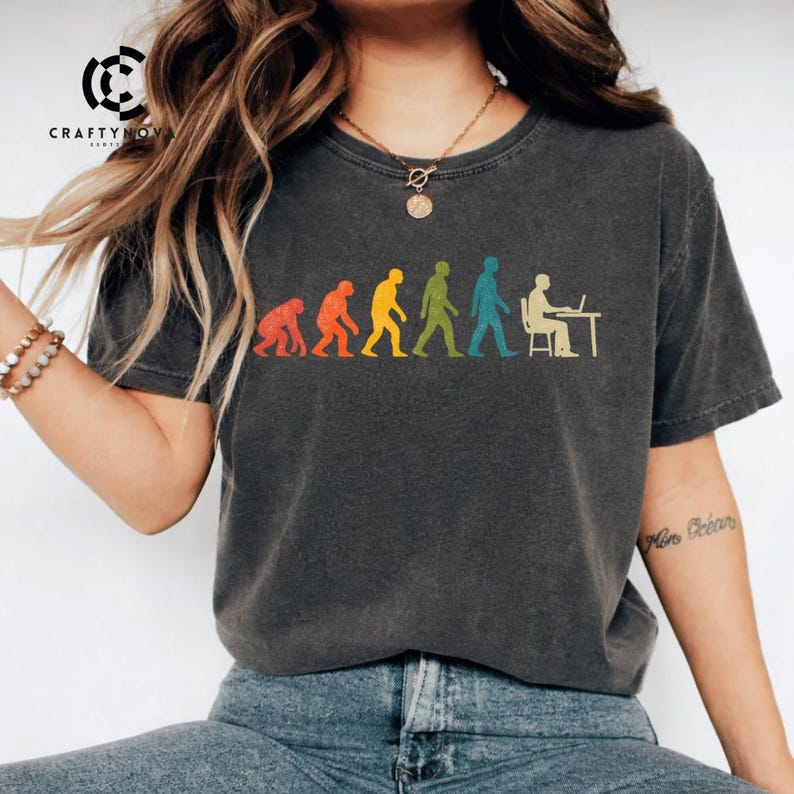 Peut inclure: Un t-shirt gris anthracite &agrave; manches courtes avec un graphique color&eacute; repr&eacute;sentant l'&eacute;volution humaine. Le motif montre une progression d'un primate vers un humain assis &agrave; un bureau avec un ordinateur, dans des tons rouge, orange, jaune, vert et bleu.