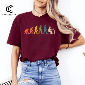 Peut inclure: Un t-shirt &agrave; manches courtes bordeaux avec une impression graphique de l'&eacute;volution humaine en orange, jaune, vert et bleu. Le motif montre la progression d'un primate vers un humain assis &agrave; un bureau avec un ordinateur, port&eacute; avec un jean bleu.