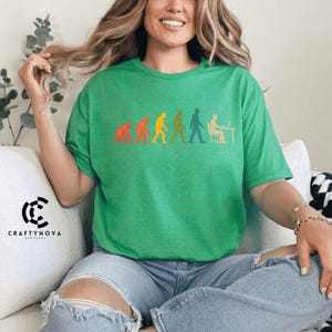 Peut inclure: Un t-shirt vert vif &agrave; manches courtes avec un imprim&eacute; graphique multicolore repr&eacute;sentant l'&eacute;volution de l'humanit&eacute;, partant d'un primate et progressant par &eacute;tapes jusqu'&agrave; une personne assise &agrave; un bureau travaillant sur un ordinateur.