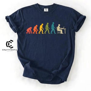 Peut inclure: Un t-shirt bleu marine &agrave; manches courtes avec un graphique multicolore repr&eacute;sentant l'&eacute;volution humaine, passant d'un primate &agrave; une personne assise &agrave; un bureau avec un ordinateur. Le design utilise des silhouettes rouges, orange, jaunes, vertes et bleues.