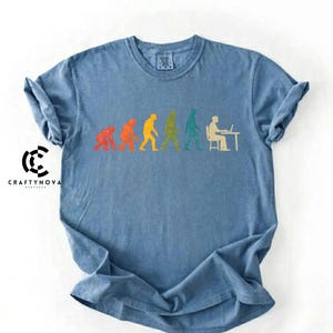Peut inclure: Un t-shirt bleu &agrave; col rond arborant un graphique color&eacute; de l'&eacute;volution humaine, commen&ccedil;ant par un primate et progressant vers un humain assis &agrave; un bureau avec un ordinateur. Le motif utilise des silhouettes rouges, orange, jaunes, vertes et bleu sarcelle.
