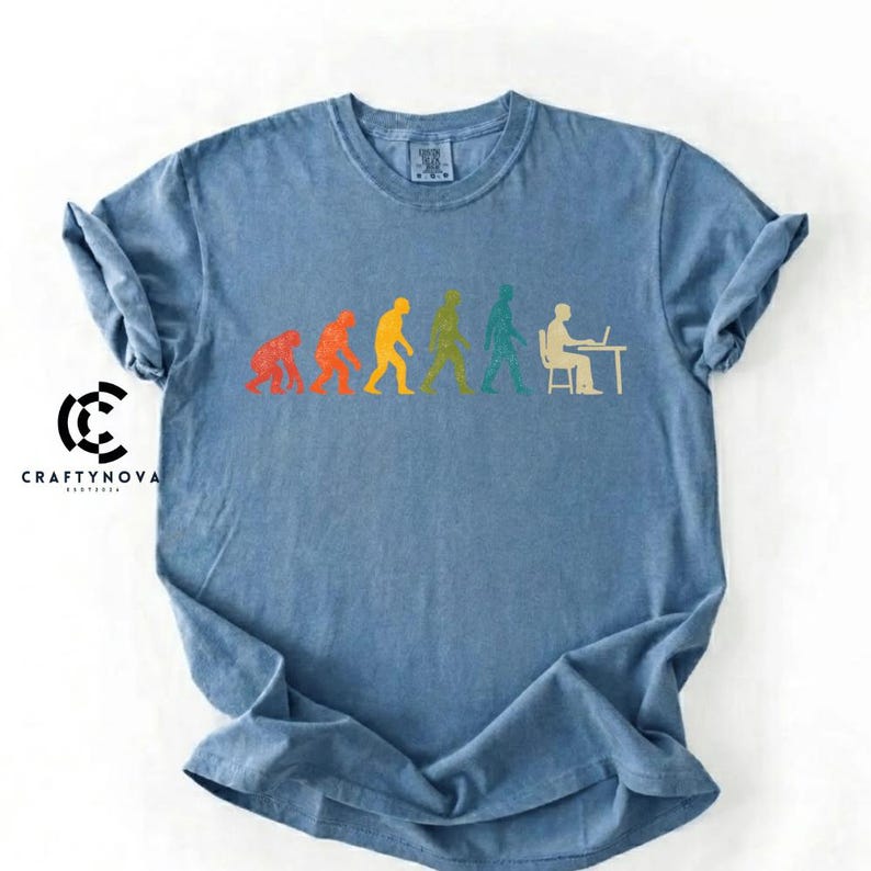Peut inclure: Un t-shirt bleu &agrave; col rond arborant un graphique color&eacute; de l'&eacute;volution humaine, commen&ccedil;ant par un primate et progressant vers un humain assis &agrave; un bureau avec un ordinateur. Le motif utilise des silhouettes rouges, orange, jaunes, vertes et bleu sarcelle.