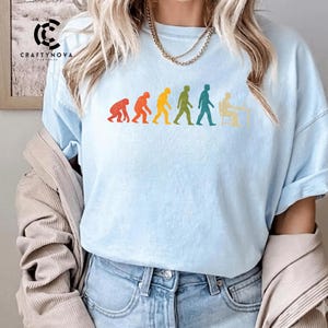 Peut inclure: Un t-shirt bleu clair avec un graphique multicolore repr&eacute;sentant l'&eacute;volution humaine, passant d'un primate &agrave; une personne assise &agrave; un bureau. Le motif utilise des silhouettes rouges, orange, jaunes, vertes et bleu sarcelle pour les &eacute;tapes.