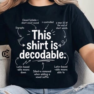 Op de afbeelding: Een zwart T-shirt met korte mouwen en witte tekst die luidt: "This shirt is decodable." Het ontwerp bevat diverse taalkundige annotaties met pijlen die naar specifieke woorden wijzen, waarbij concepten zoals gesloten lettergrepen, r-gecontroleerde klinkers, tweeklanken en achtervoegselregels worden uitgelegd.