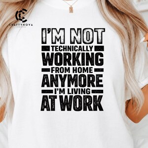 May include: A white t-shirt featuring bold, distressed black text that reads: I'M NOT TECHNICALLY WORKING FROM HOME ANYMORE I'M LIVING AT WORK. The design is centered on the chest, with a small logo in the upper left corner reading CRAFTYNOVA.