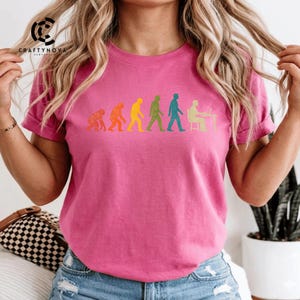 Peut inclure: Un t-shirt rose vif &agrave; manches courtes avec un imprim&eacute; graphique multicolore repr&eacute;sentant l'&eacute;volution de l'humanit&eacute;, partant d'un primate et progressant par &eacute;tapes jusqu'&agrave; une personne assise &agrave; un bureau avec un ordinateur.